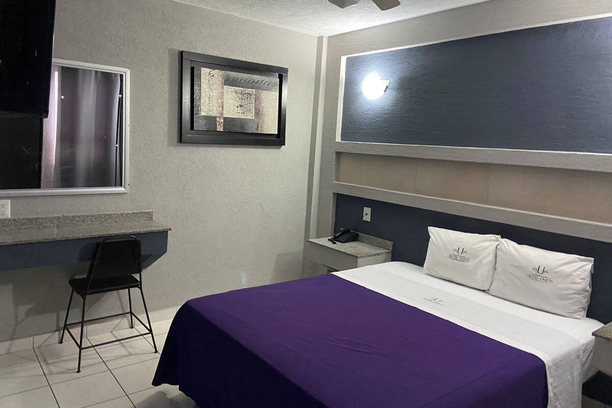 A hotel room with a bed, a desk, a chair, a ceiling fan, and a window.