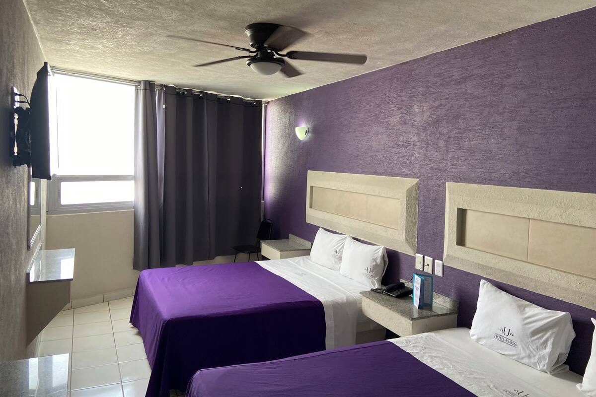 A hotel room with two beds, a ceiling fan, a window with curtains, and a wall-mounted TV.