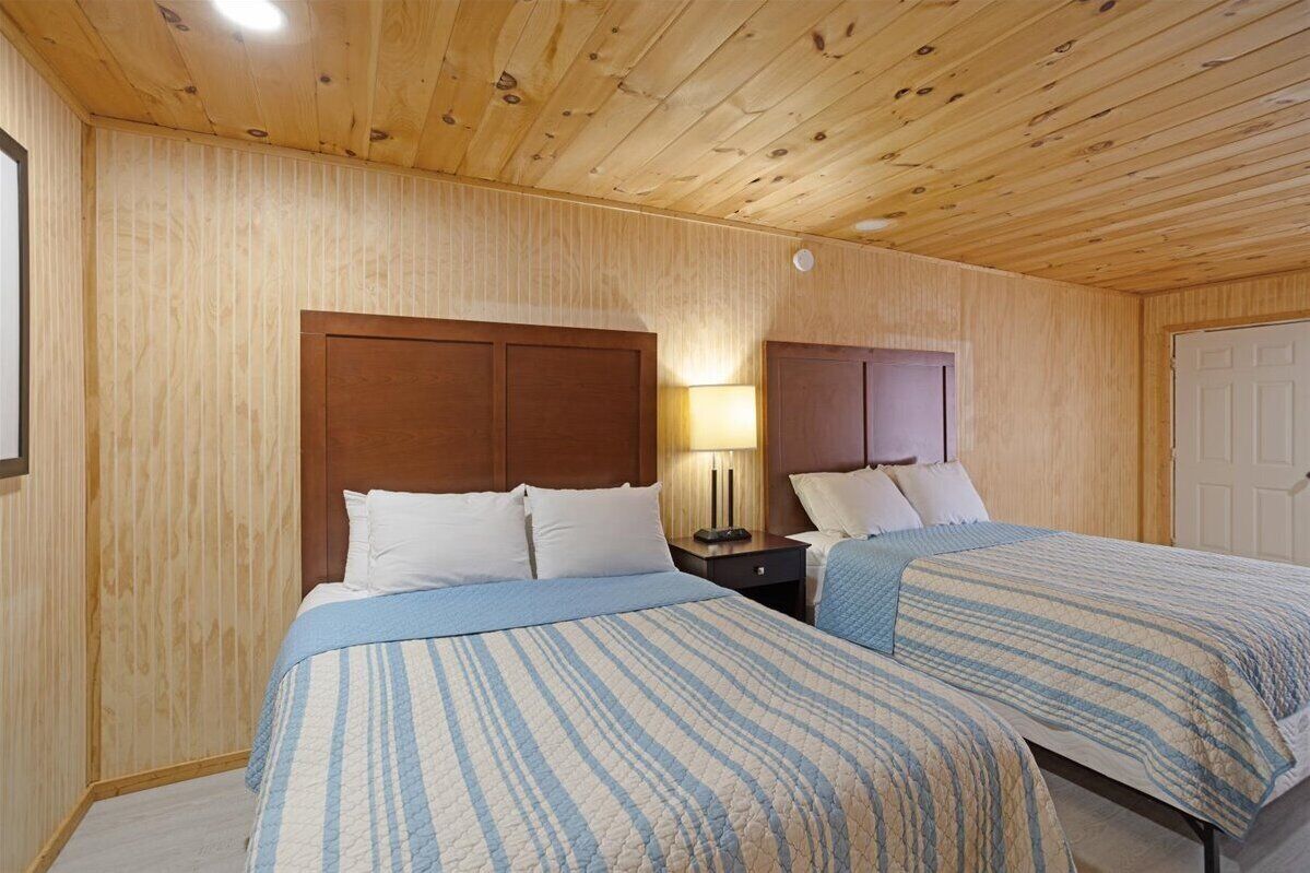 A room with two beds, wooden panel walls, a ceiling with wooden beams, and a nightstand with a lamp.