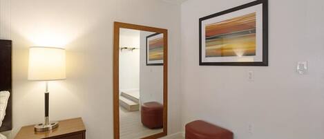Ein modernes Hotelzimmer mit einem Holznachttisch, einem Spiegel, einem roten Hocker und einem gerahmten abstrakten Gemälde an der Wand.