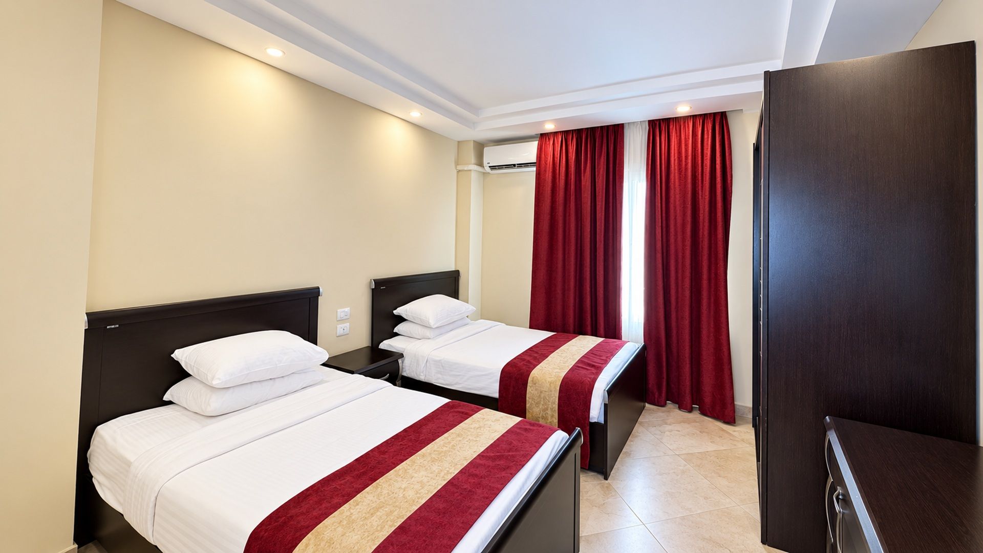 Hotellihuone, jossa on kaksi sänkyä, tumma puinen vaatekaappi ja punainen verho.
