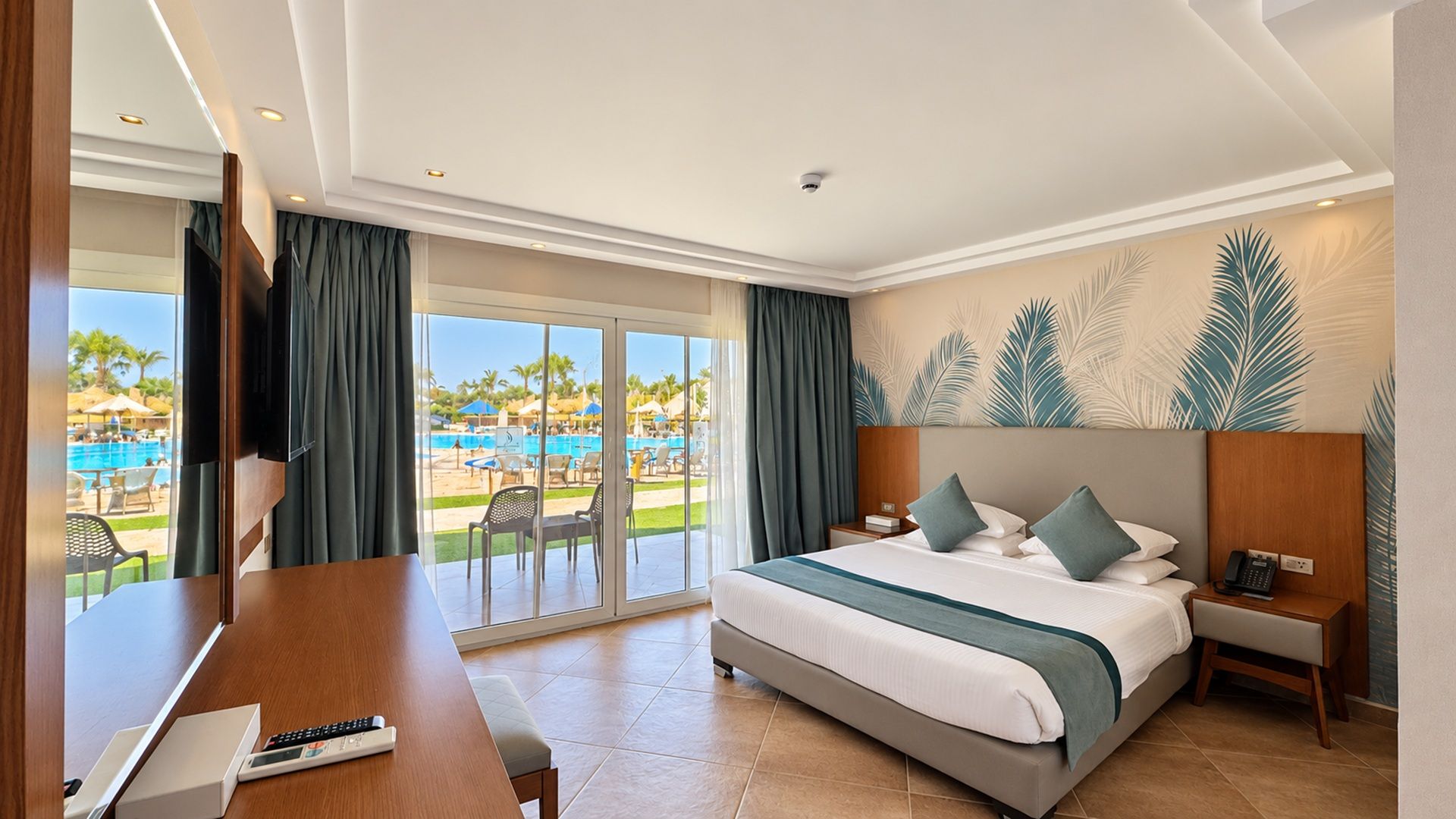 Hotelkamer met een bed, bureau, stoel, nachtkastje en uitzicht op een zwembad en palmbomen.