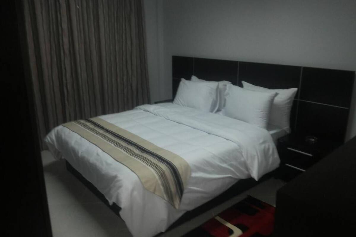 A neatly made bed with white linens and a striped throw blanket, a black headboard, and a red and black patterned rug.