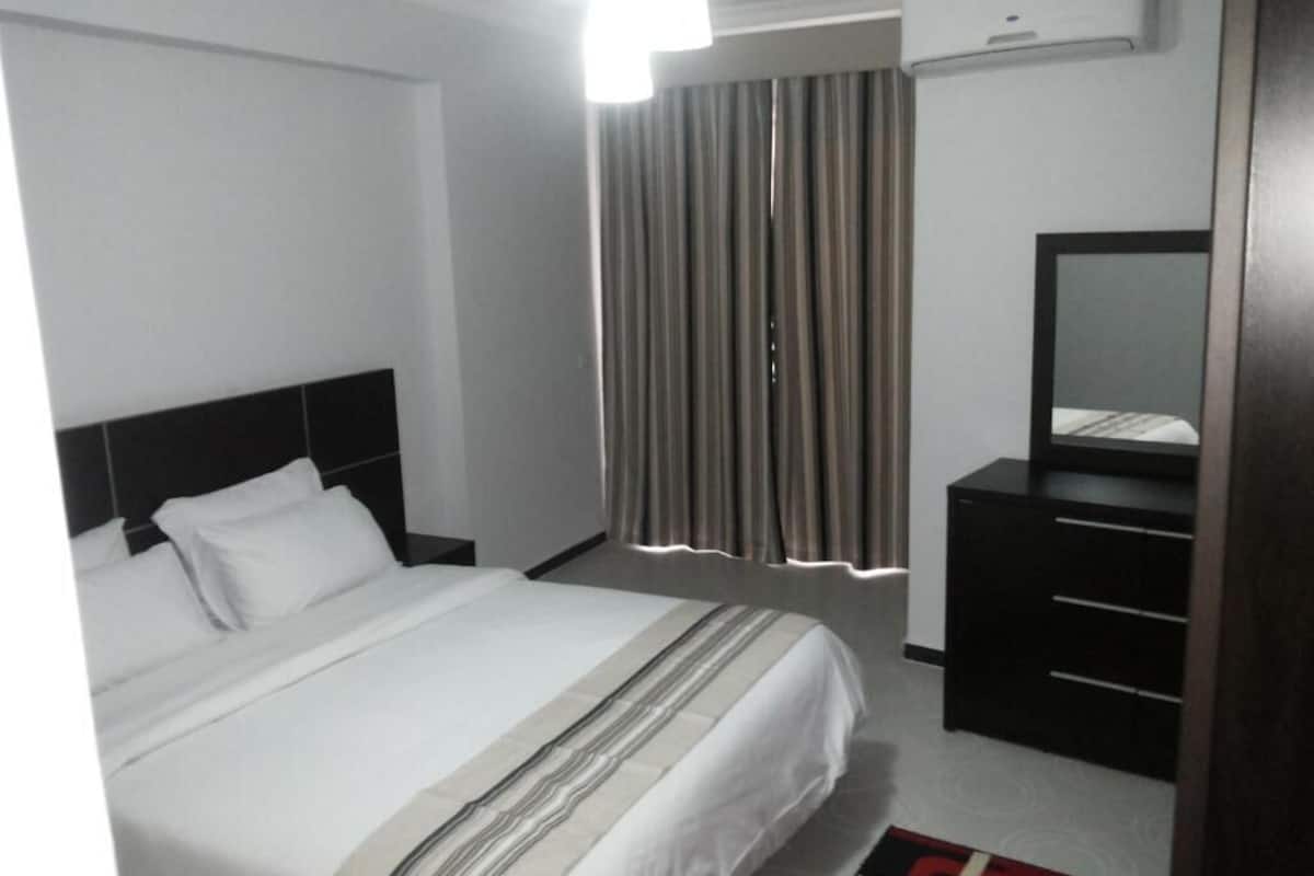 A hotel room with a bed, a dresser, a mirror, a window with curtains, and an air conditioning unit.