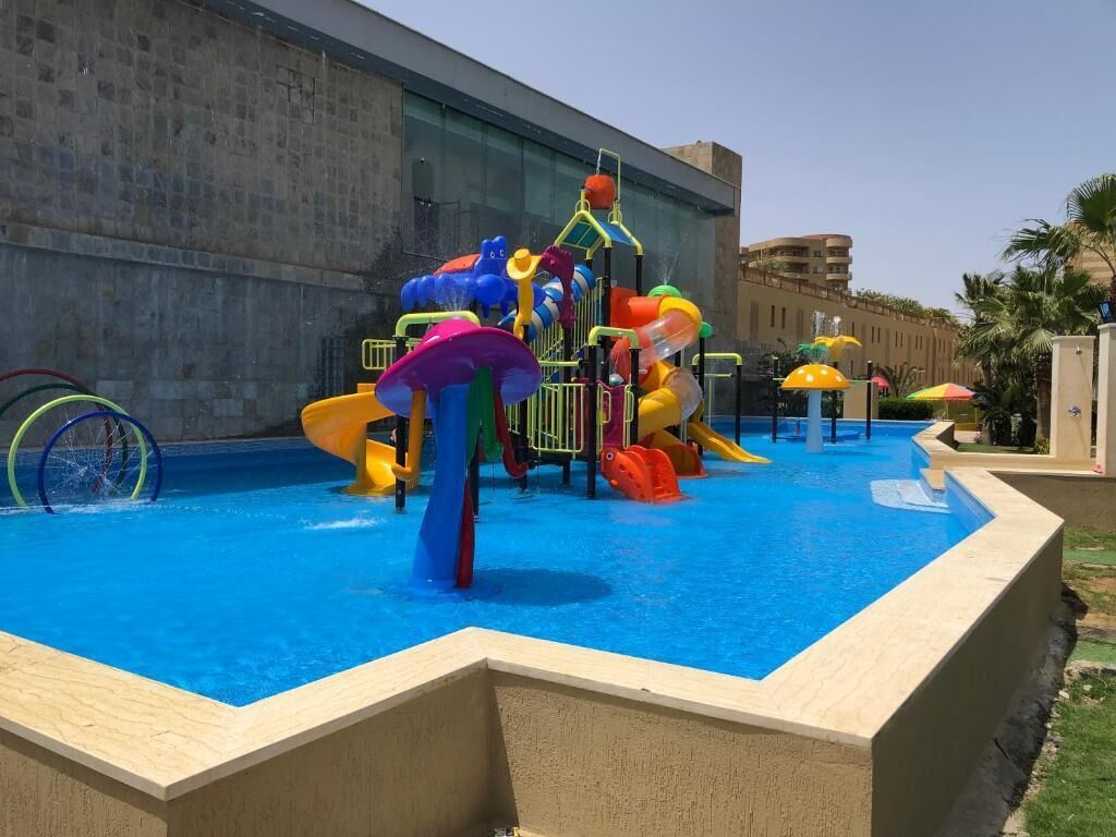 Un área de juegos acuáticos con toboganes y estructuras para escalar, rodeada por una piscina y un edificio al fondo.