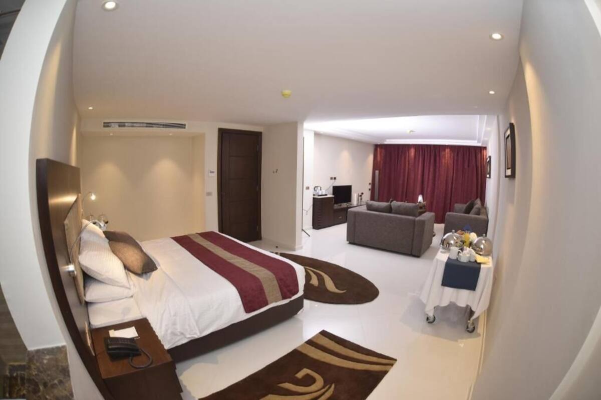 A modern hotel room with a large bed, a sofa, a small table with a tray, and a separate living area with a couch and a TV.