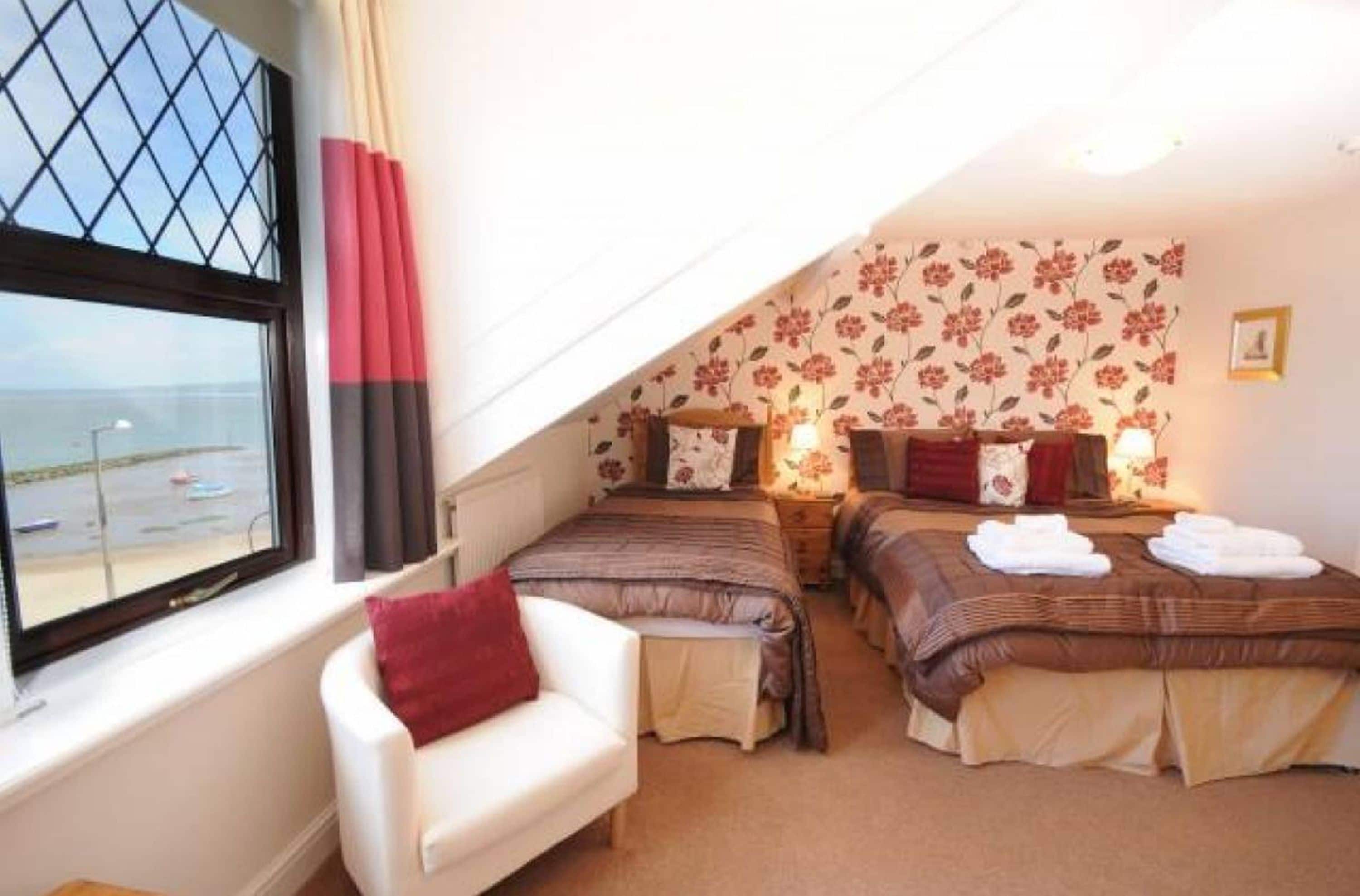 Un dormitorio con dos camas, una ventana con vista al mar y papel tapiz con motivos florales.