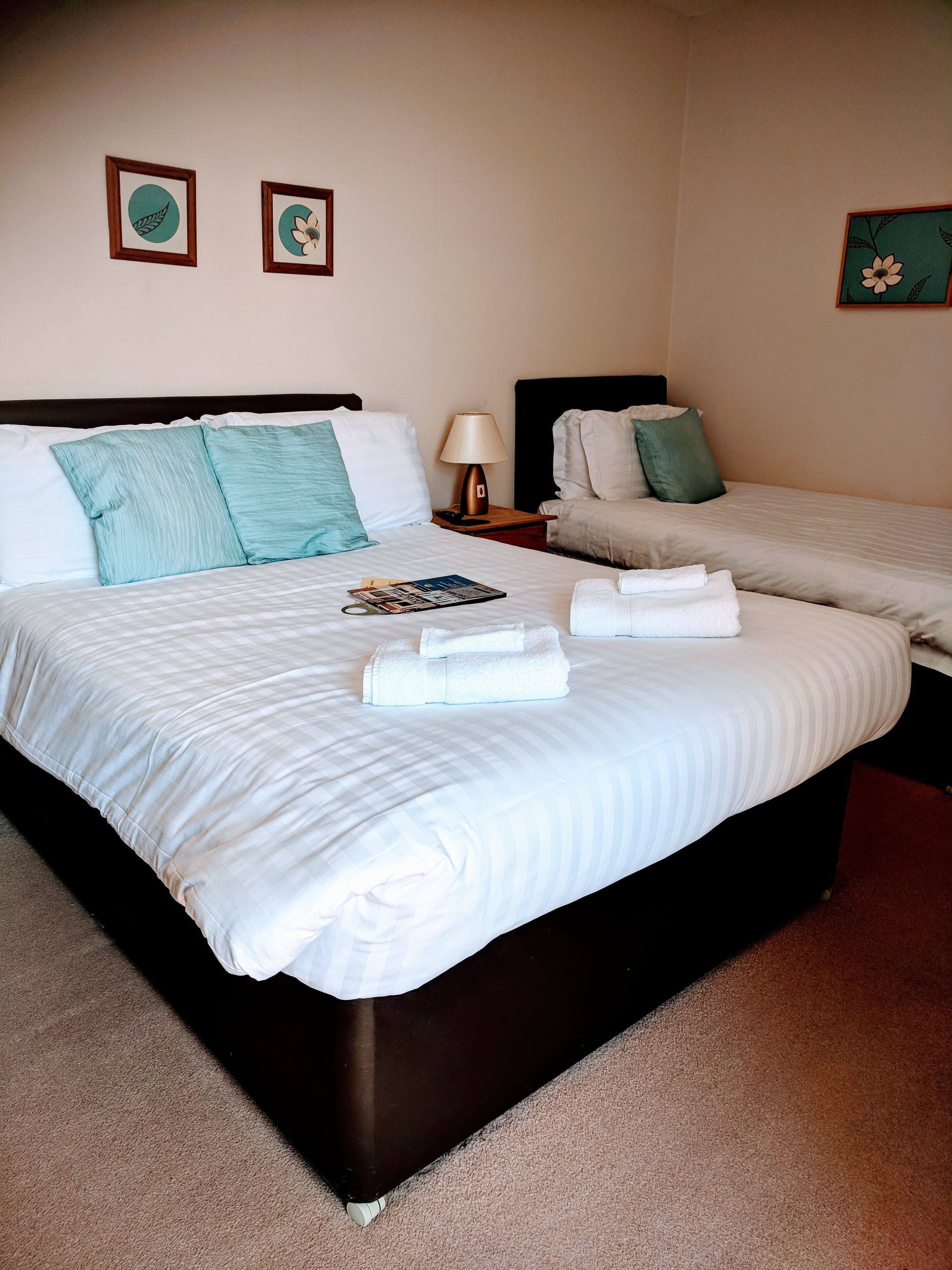 Quarto de hotel com duas camas, cada uma com roupa de cama branca e almofadas verde-água. Um criado-mudo com abajur fica entre as camas.