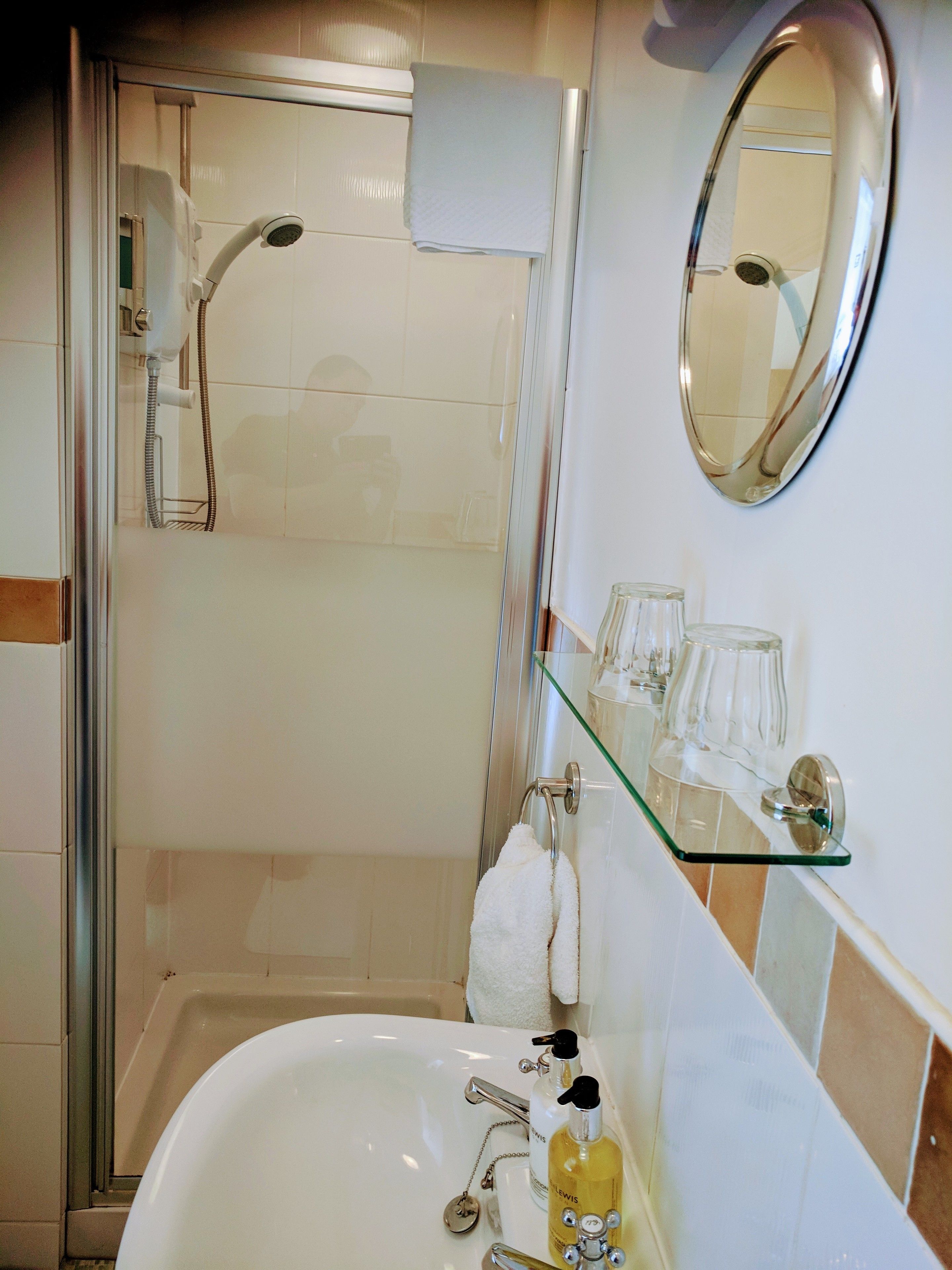 Ein Badezimmer mit Duschkabine aus Glas, einem weißen Waschbecken mit Spiegel darüber und einer Glasablage für Toilettenartikel.