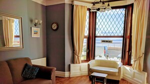 A living room with a brown sofa, a white armchair, a small wooden table, and a large window with curtains. - The Craigwell Hotel (Morecambe)