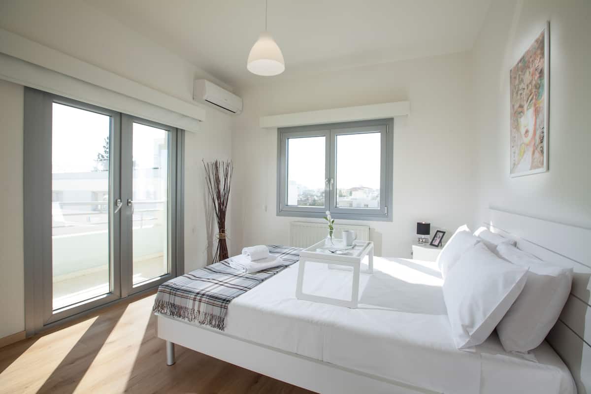 Quarto moderno com cama grande, mesa de cabeceira, janela e vista para a cidade.