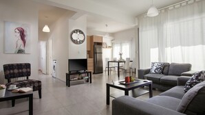 A modern living room with a sofa, armchair, coffee table, and a TV. There is a dining area with a table and chairs, and a kitchen with a refrigerator and microwave.
