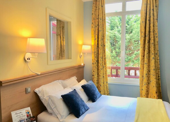 A hotel room with a bed, two wall-mounted lamps, a mirror, and a window with curtains.