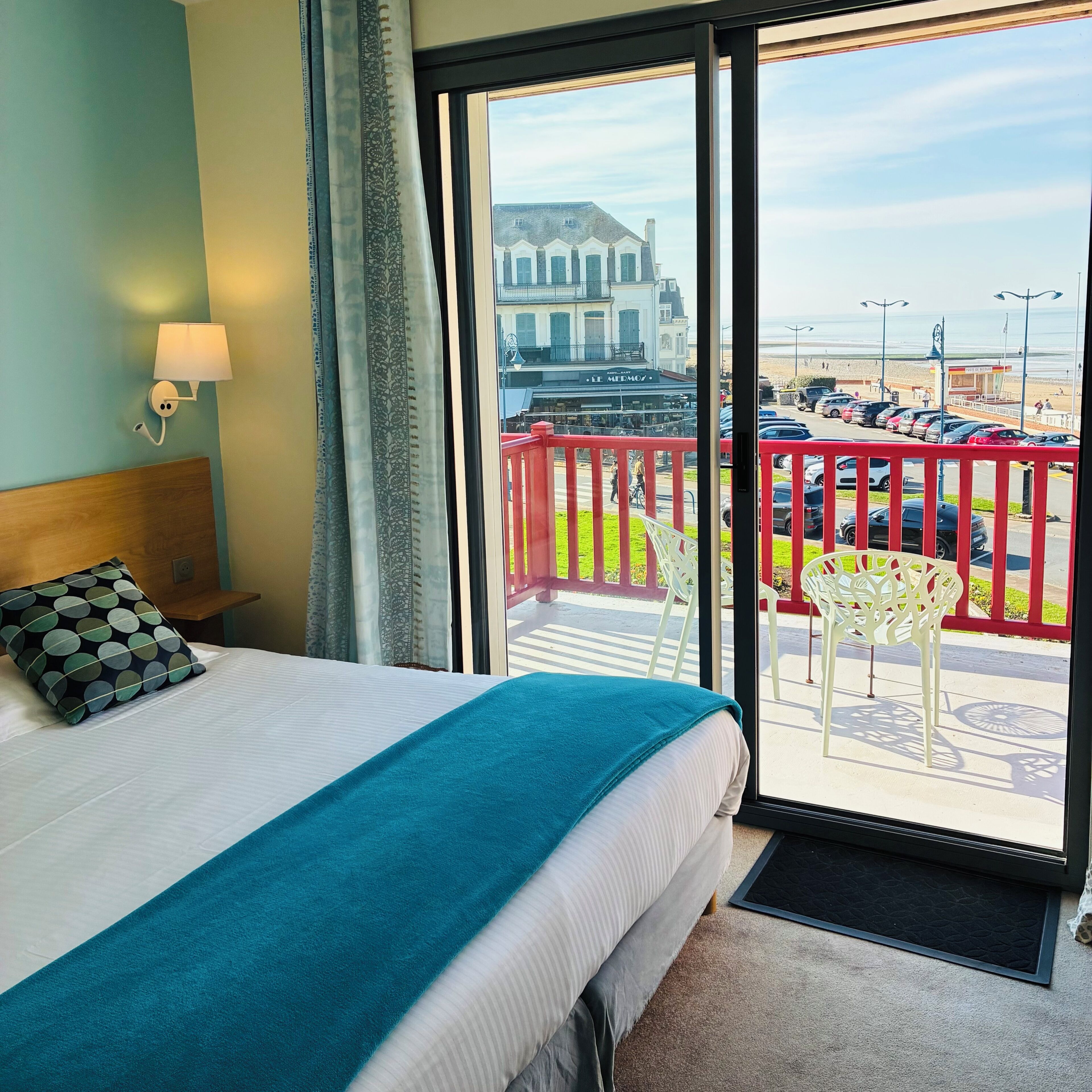 A hotel room with a bed, a view of the beach, and a balcony with chairs.