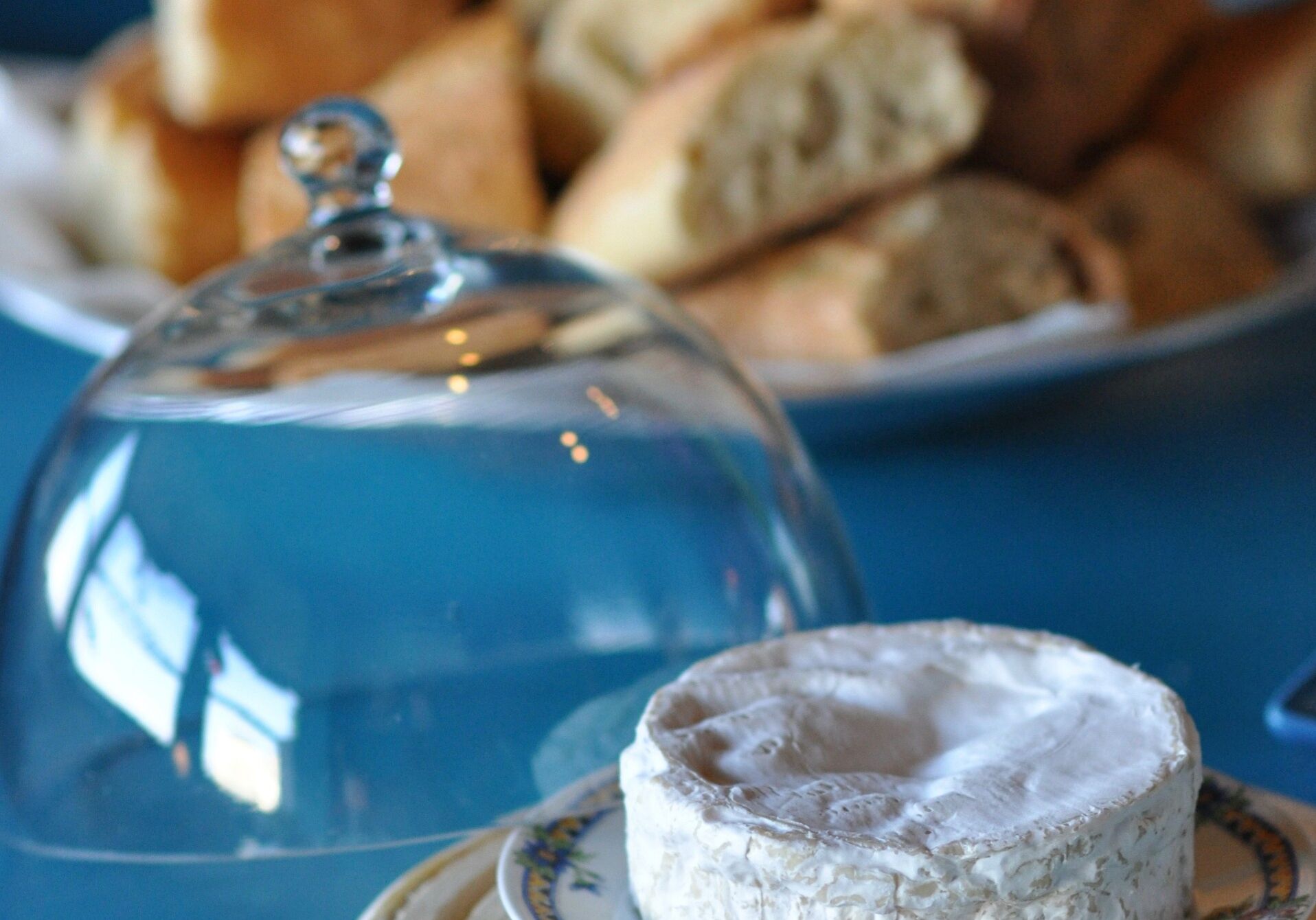 A cheese wheel under a glass dome on a table with a plate of bread in the background.