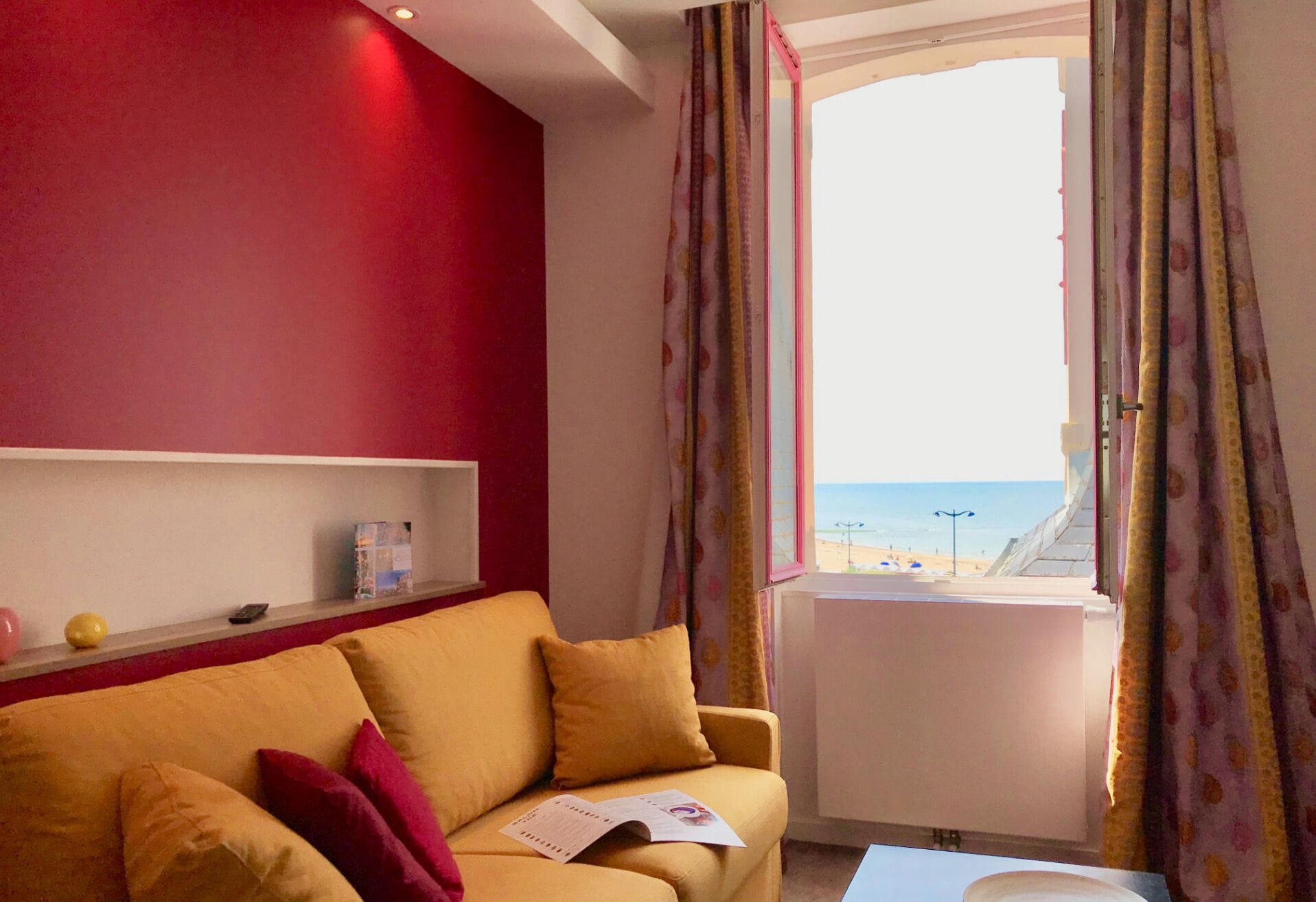 Ein Zimmer mit roter Wand, einem gelben Sofa mit roten Kissen, einem kleinen Regal mit Buch und Flasche sowie einem Fenster mit Blick auf den Strand und das Meer.