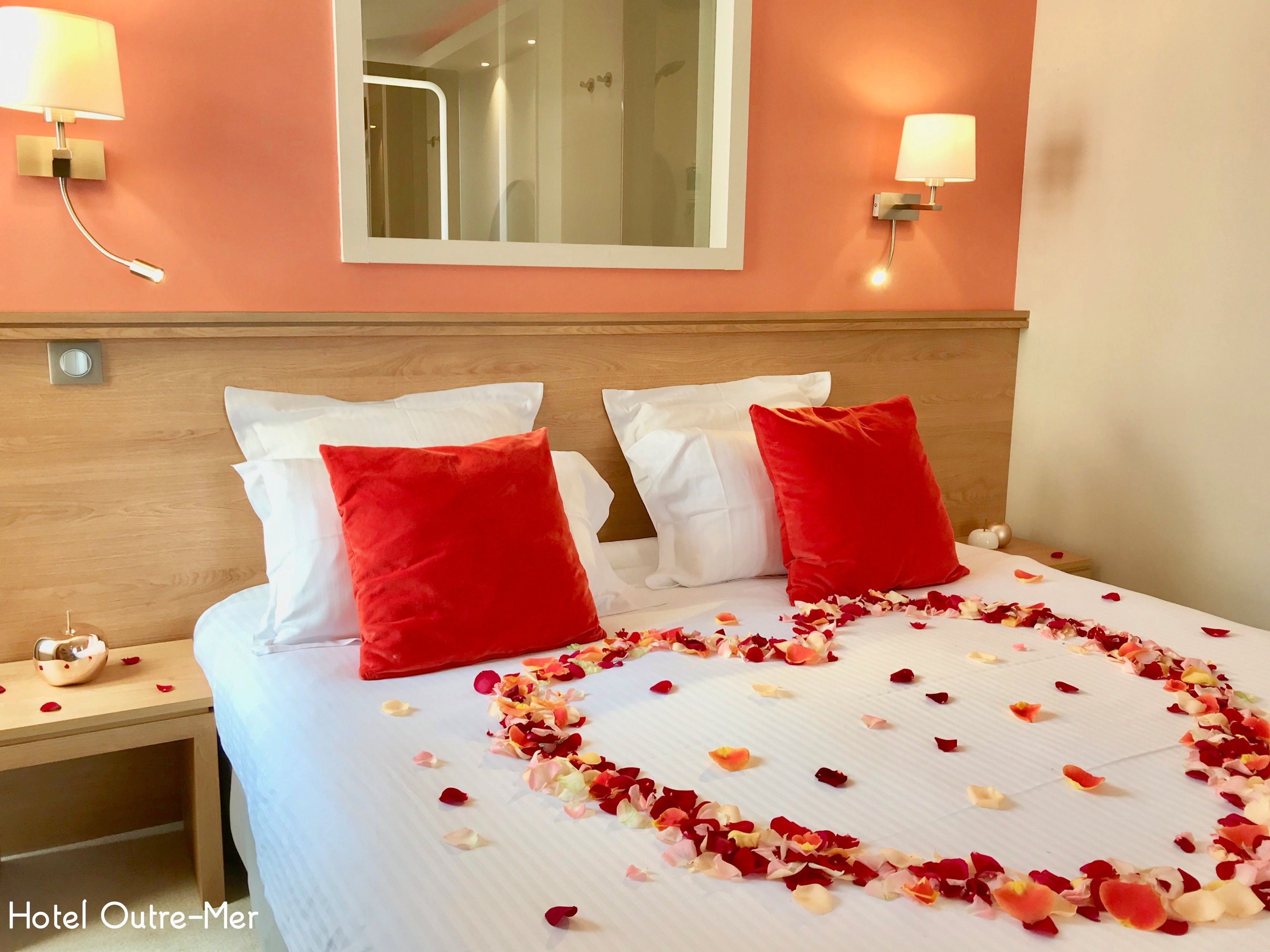 A hotel room with a bed, two red pillows, a bedside table with a lamp, and a mirror on the wall.