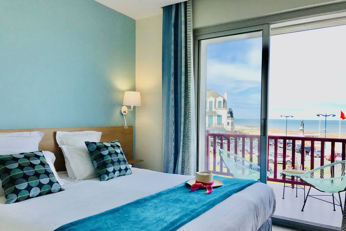 A hotel room with a large bed, a view of the beach, and a balcony with chairs.
