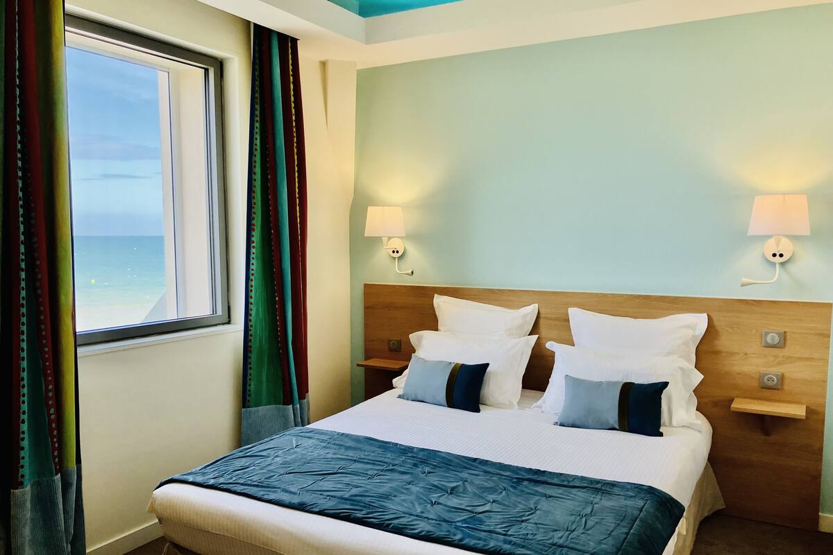 A hotel room with a bed, two bedside tables, a window with curtains, and a view of the sea.