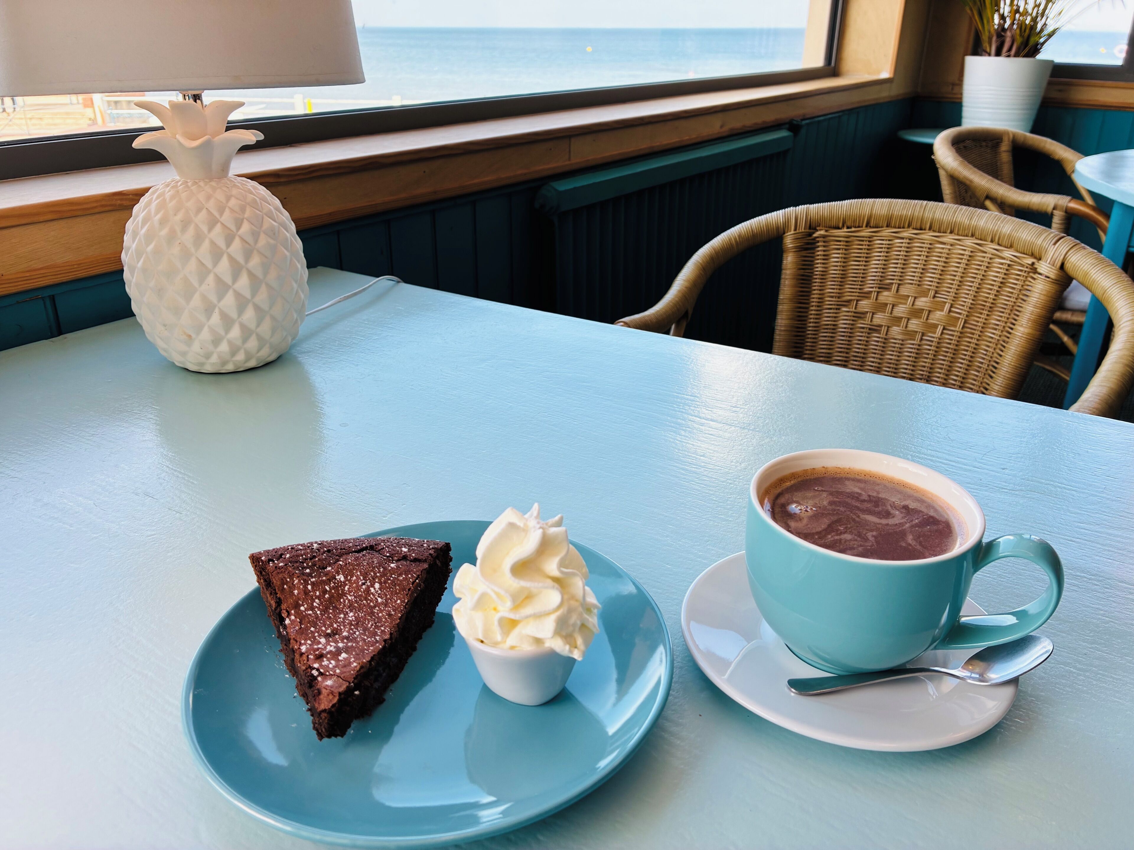 Una mesa con una rebanada de pastel y crema batida, una taza de chocolate caliente y vista al océano a través de una ventana.