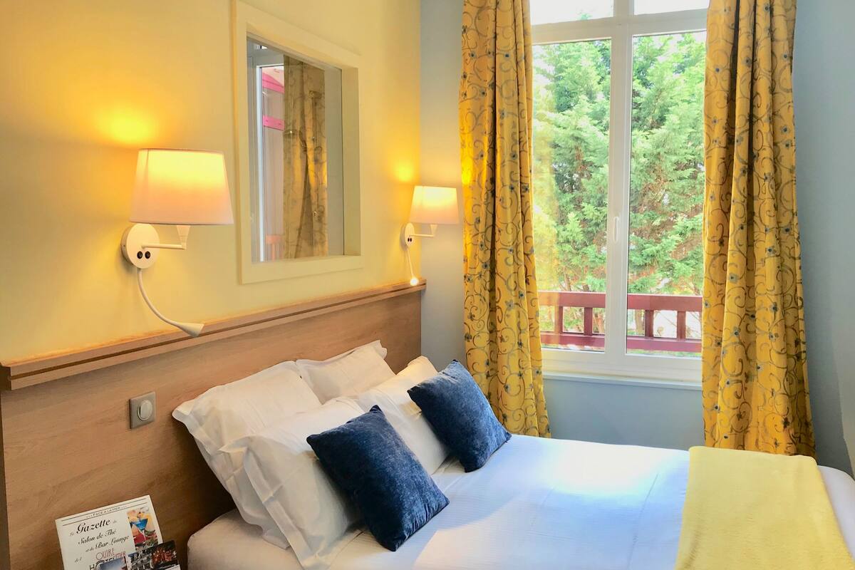 A hotel room with a bed, two wall-mounted lamps, a mirror, and a window with curtains.