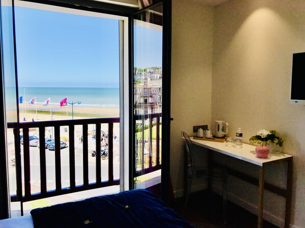 A room with a balcony overlooking a beach, a small table with a chair, and a bed. - Hôtel Outre-Mer (Villers-sur-Mer)