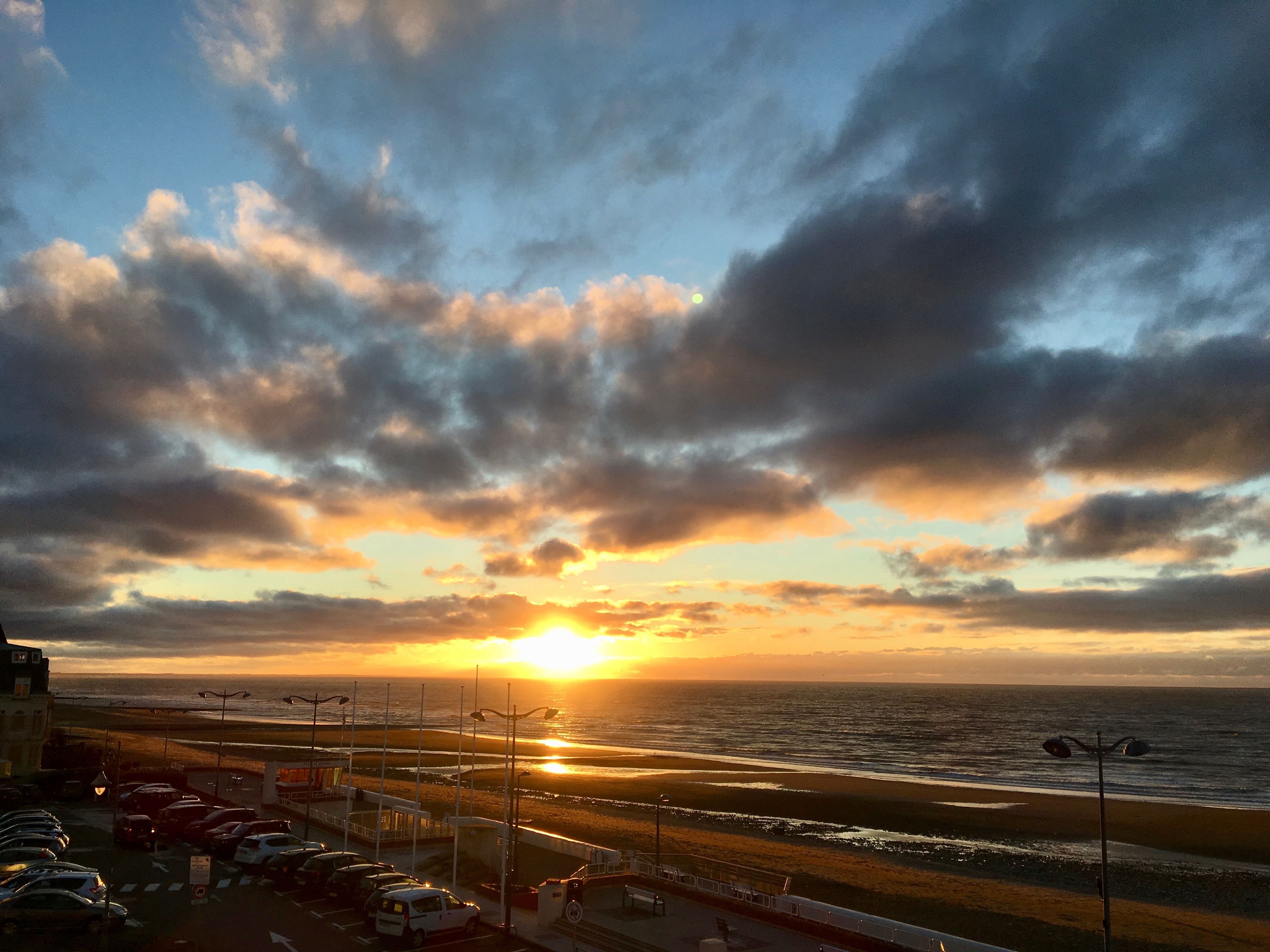 A sunset view over a beach with parked cars and a road in the foreground.