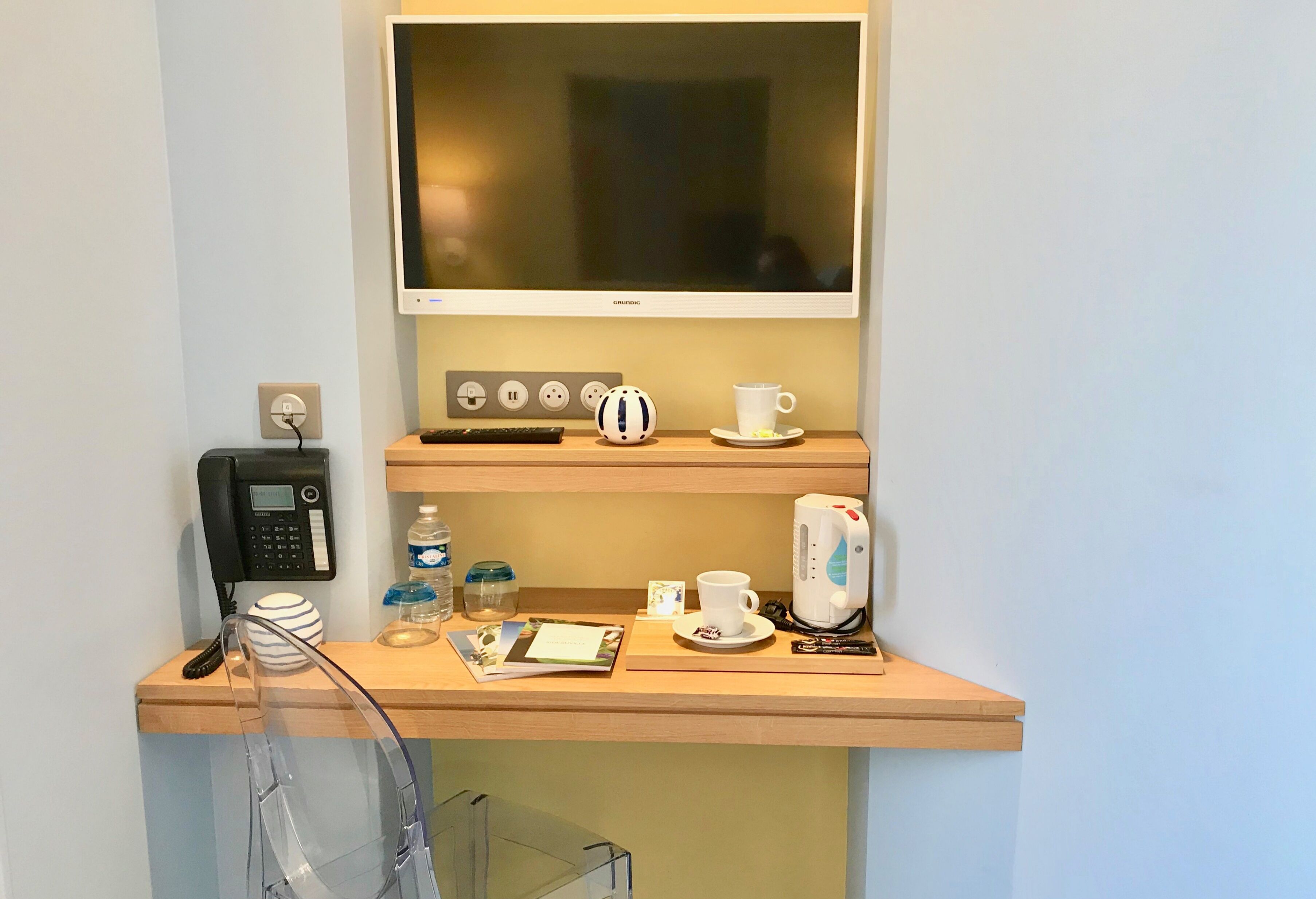 A hotel room with a mounted TV, a desk with a phone, a kettle, a cup, and a water bottle.