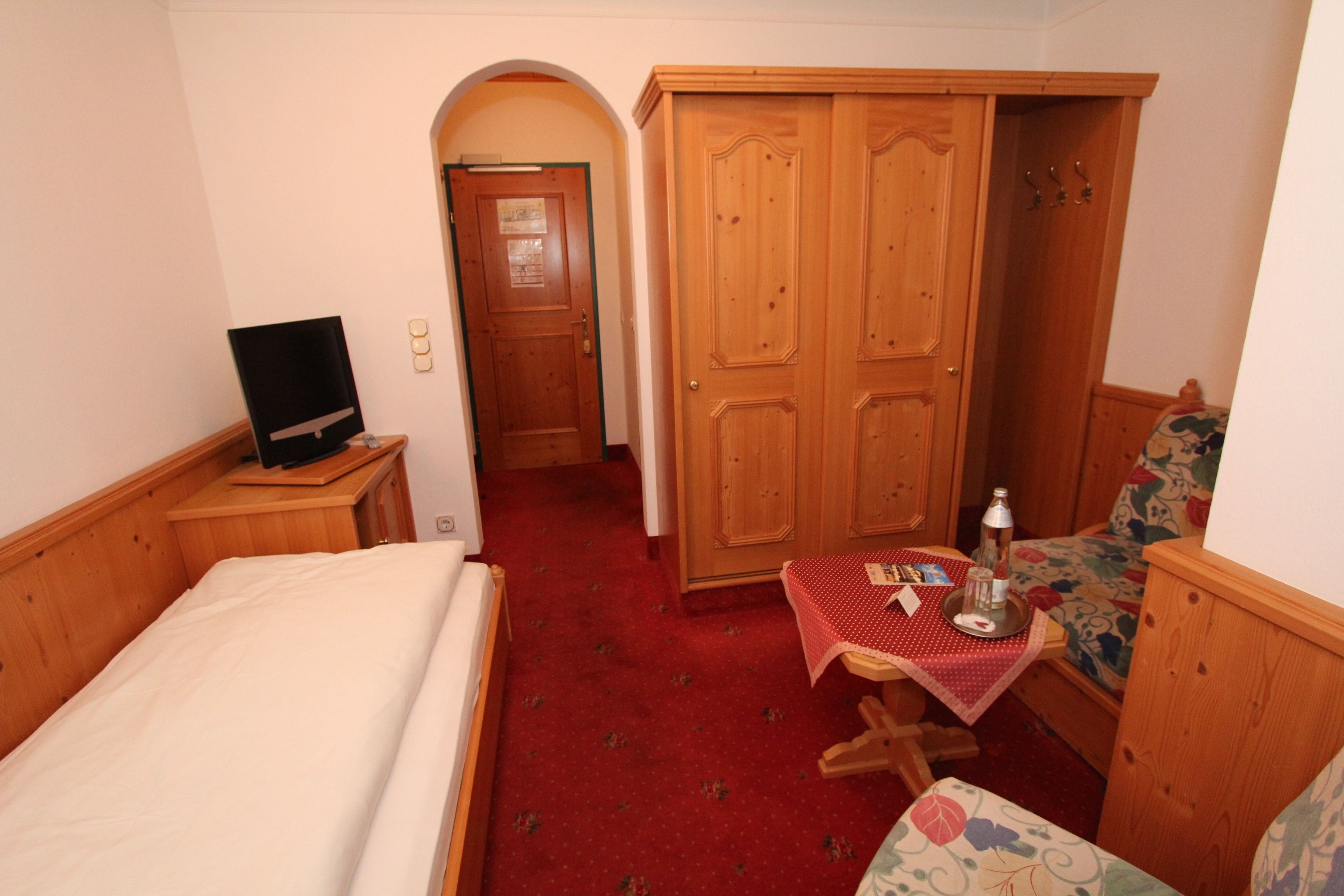 A hotel room with a bed, a television on a wooden nightstand, a small table with a red tablecloth, and a wooden wardrobe.