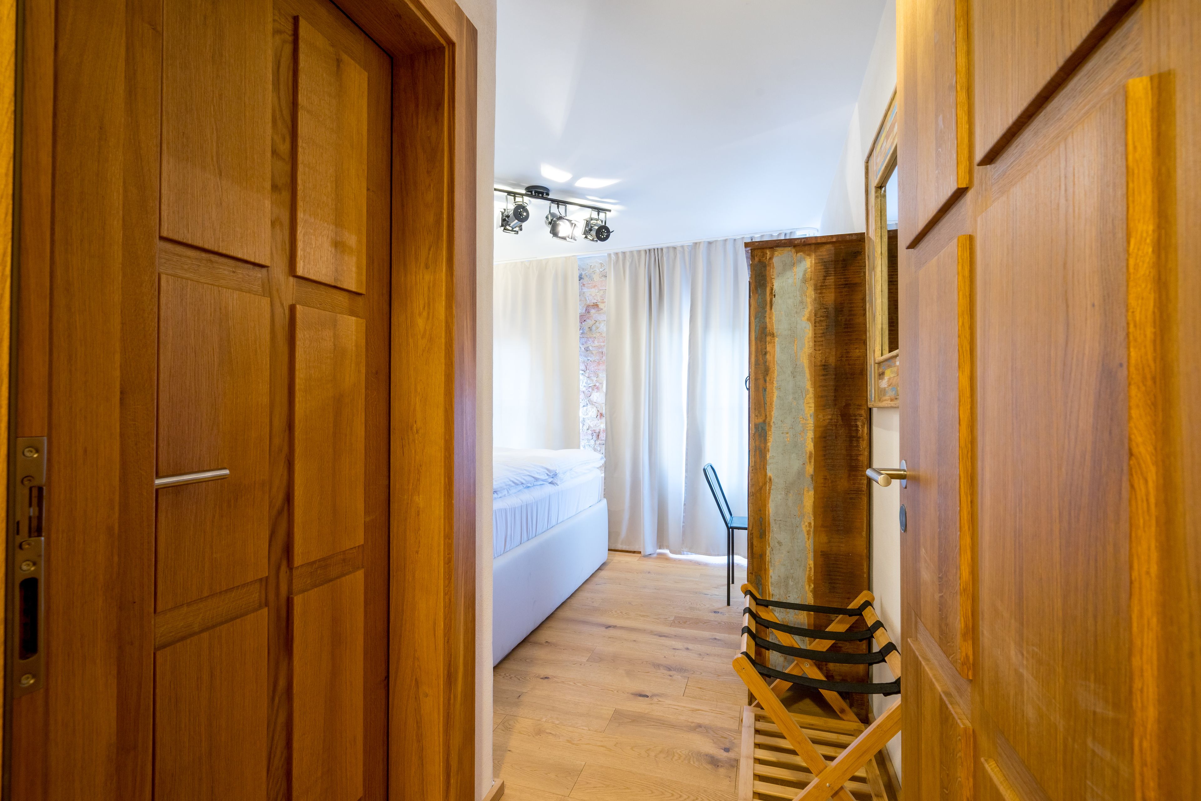 A wooden hallway leading to a bedroom with a bed, curtains, and a staircase.