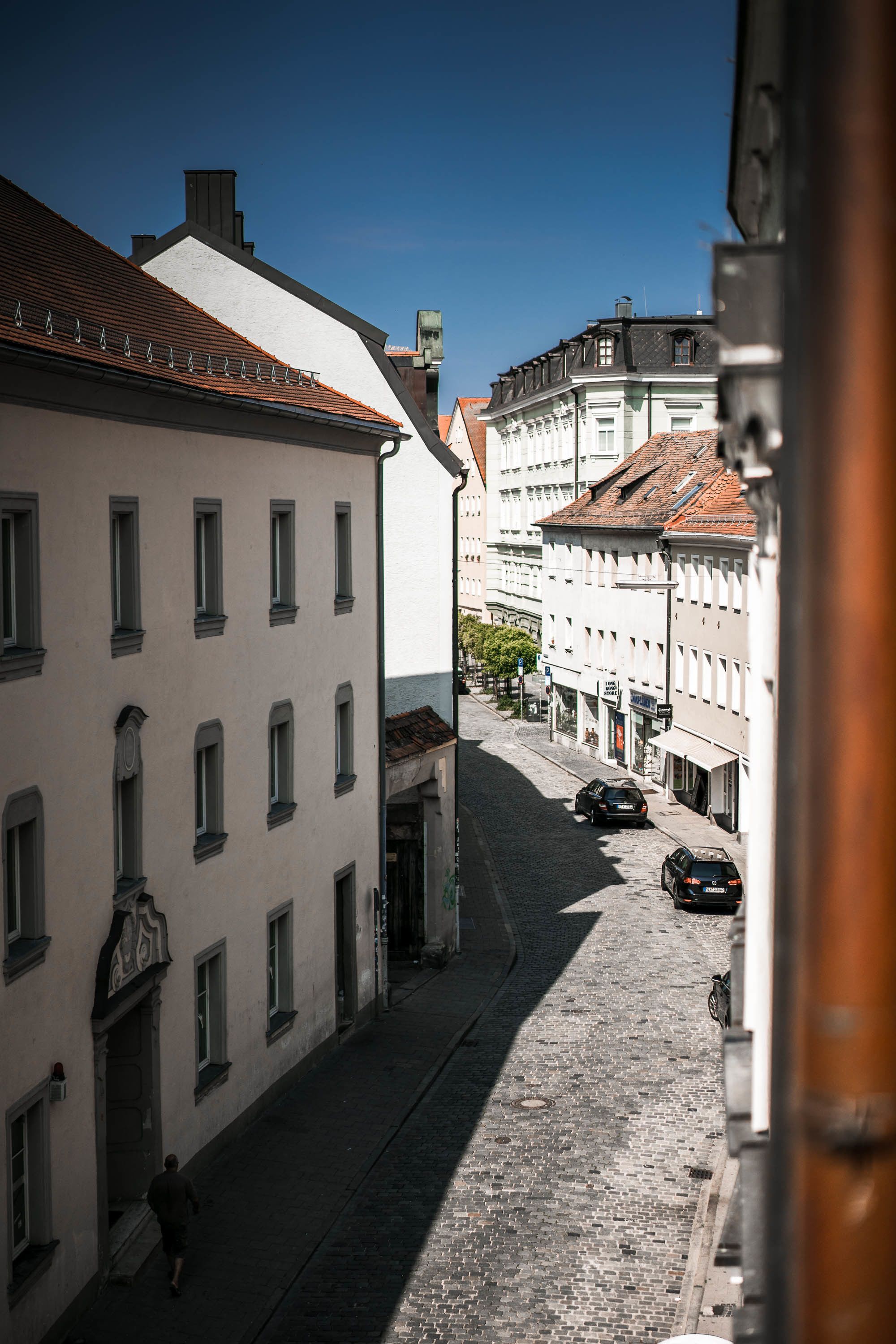 Rua de paralelepípedos estreita, ladeada por edifícios históricos, com uma pessoa caminhando e carros estacionados.