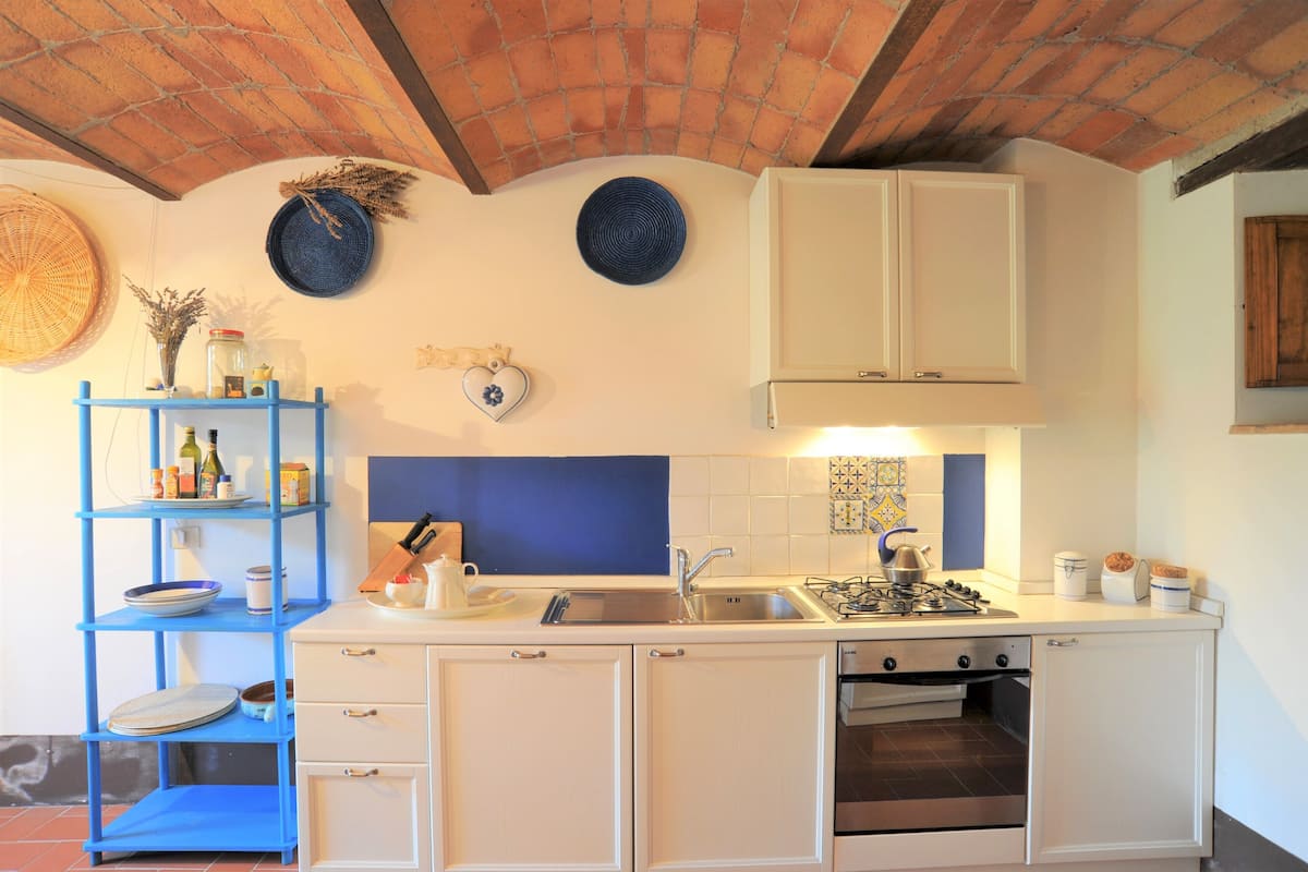 Een kleine keuken met witte kastjes, een blauwe plank, een gasfornuis en een betegelde spatwand.