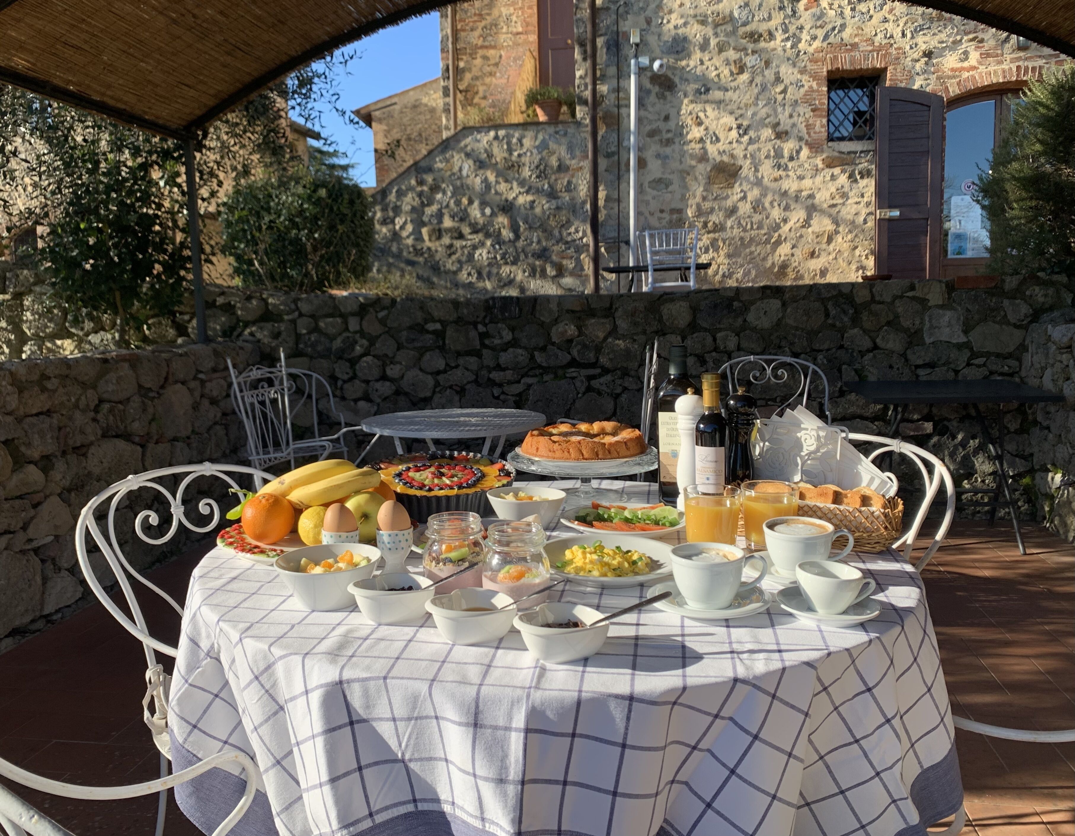An outdoor dining area with a stone wall backdrop, a table set with breakfast items, and a view of a rustic stone building.