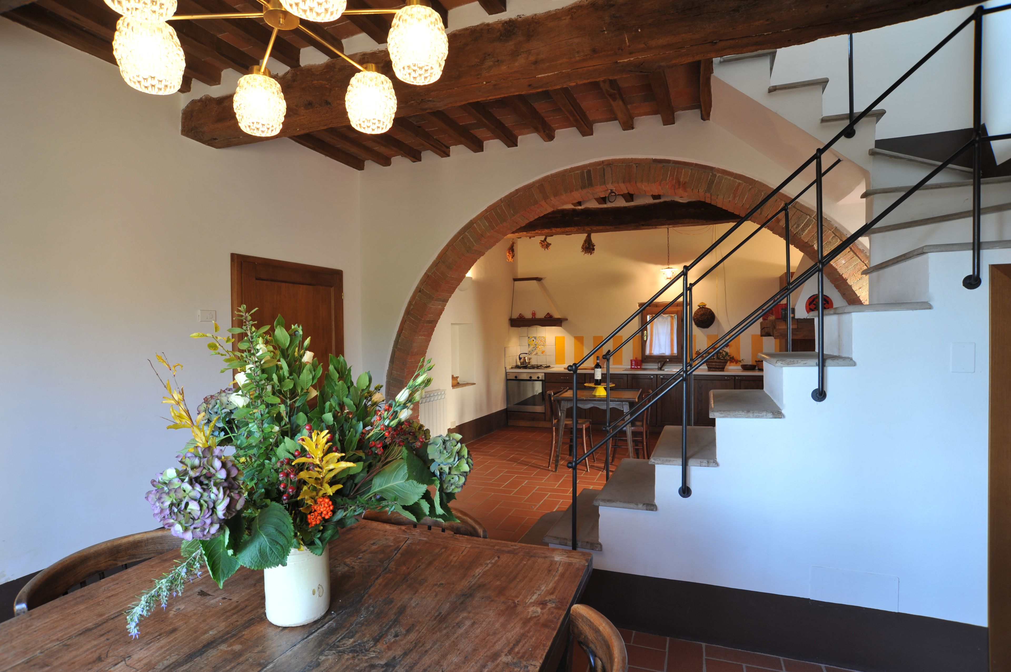 Un área de comedor con una mesa de madera y un centro de mesa floral, un arco que da a una cocina y una escalera con barandilla de metal.