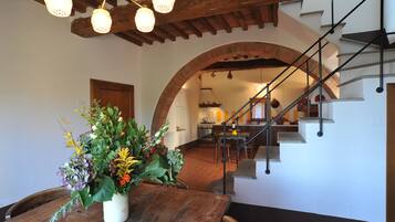 Un área de comedor con una mesa de madera y un centro de mesa floral, un arco que da a una cocina y una escalera con barandilla de metal.