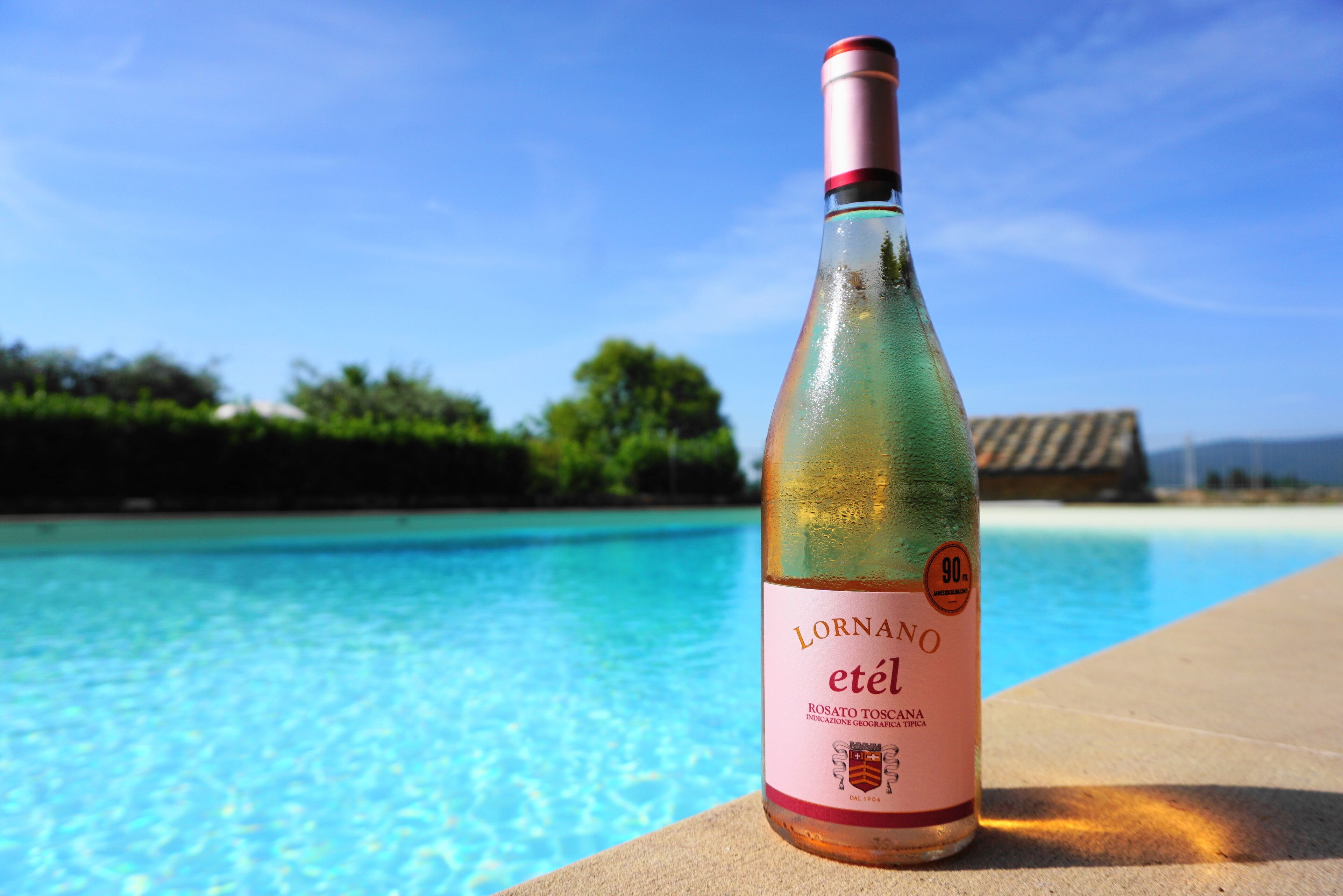 Eine Flasche Lornano etel Wein mit Etikett, platziert am Pool mit klarem, blauem Wasser und Blick auf Strand und Bäume im Hintergrund.