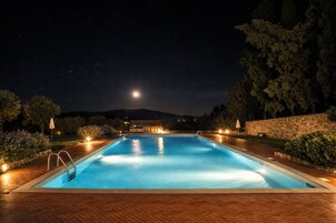 Una piscina rectangular de noche con cubierta de madera, rodeada de árboles y un muro de piedra.