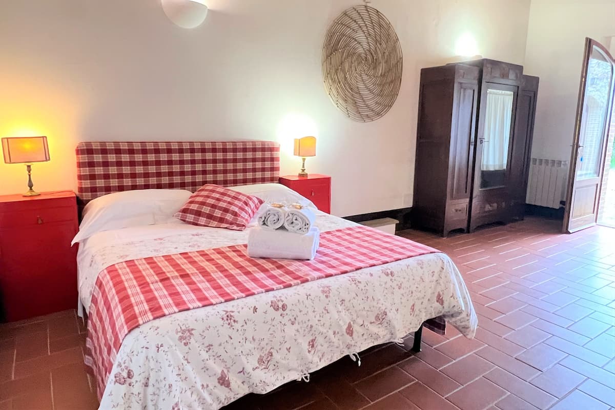 Een slaapkamer met een geruit beddegoed, een opgerolde handdoek op het bed, een rode nachtkastje, een wandventilator en een kledingkast.