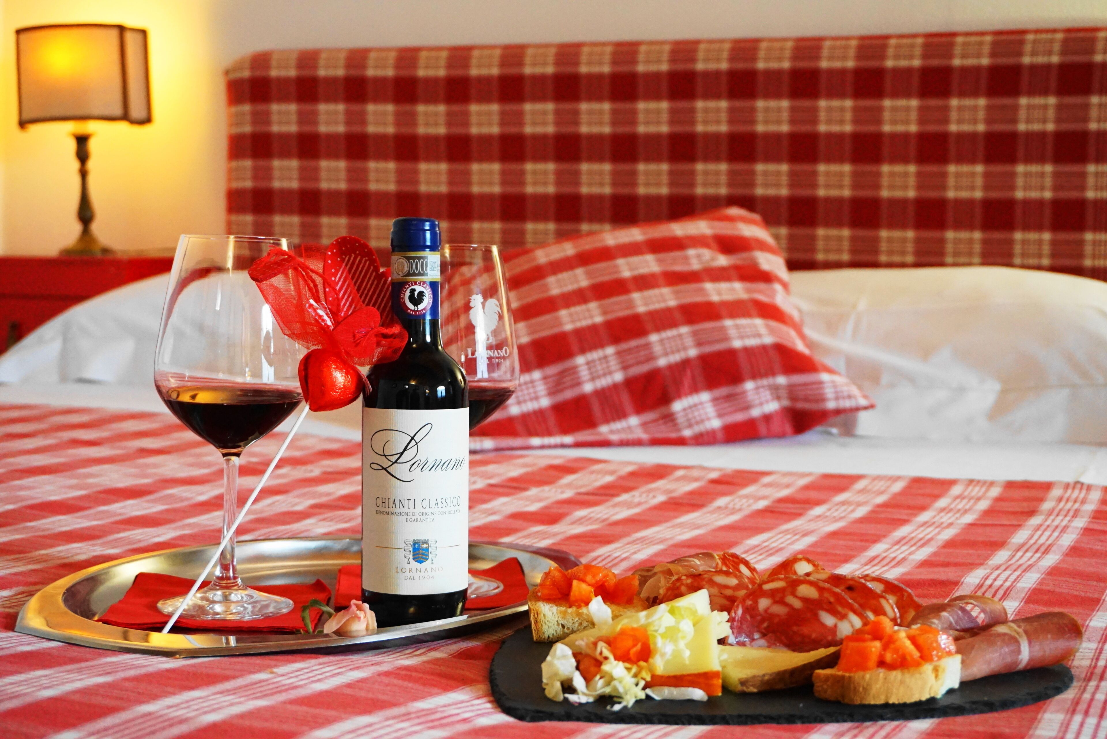 A bed with a red and white checkered bedspread, a bottle of Chianti Classico wine, a glass of red wine, and a tray with various cheeses, meats, and bread.