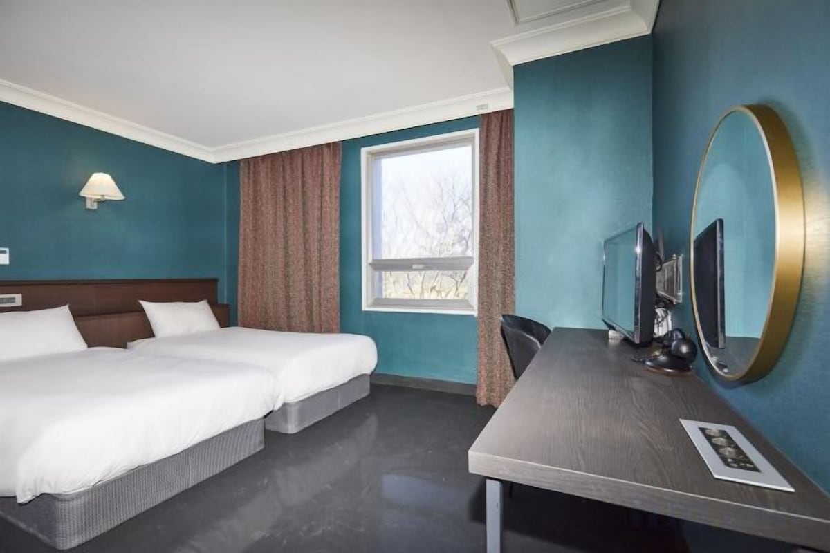 A hotel room with two beds, a desk, a chair, a window with curtains, and a mirror.
