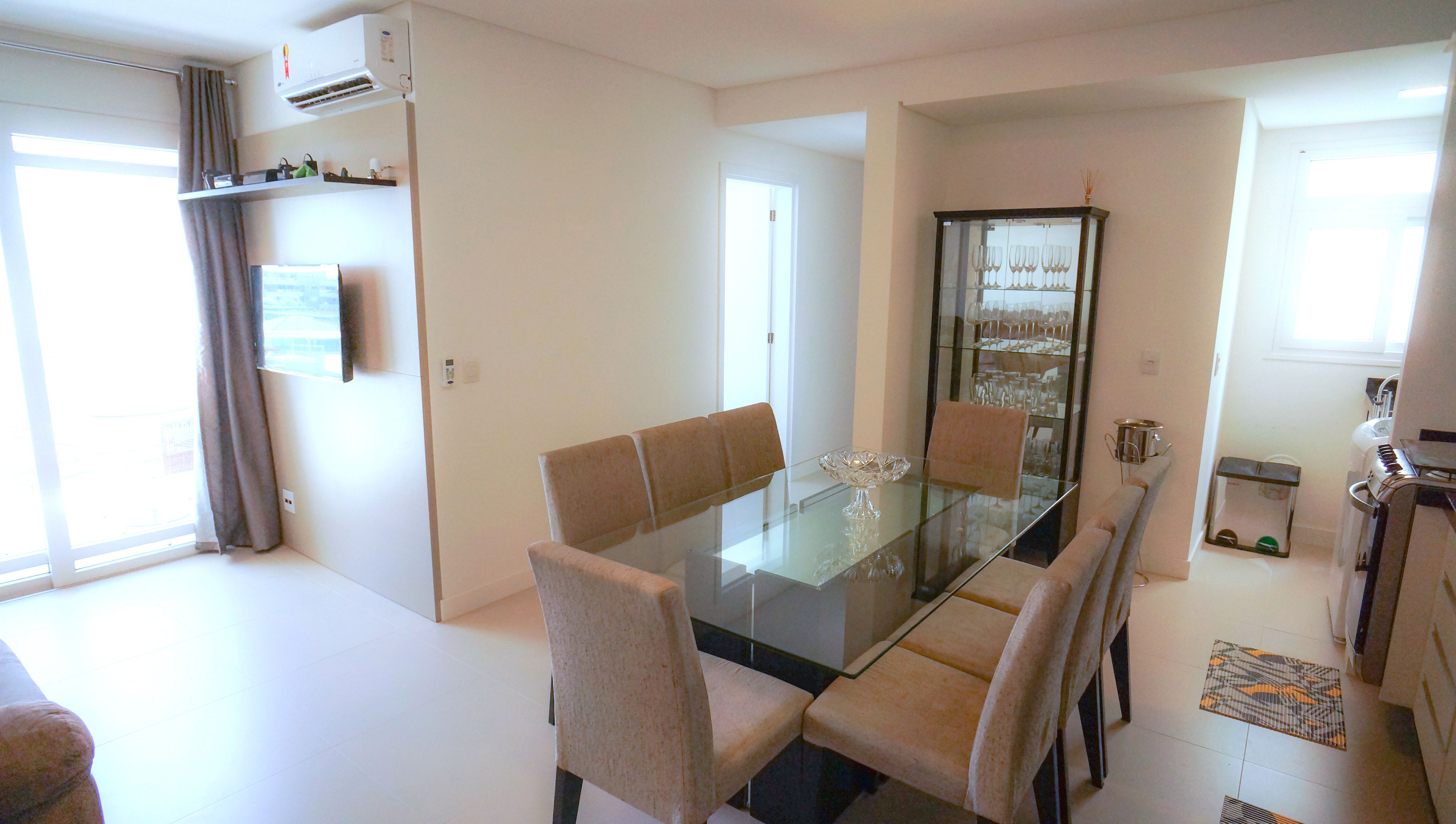 A modern dining area with a glass table and cushioned chairs, a glass cabinet with wine glasses, and a kitchen area with a kettle and sink.