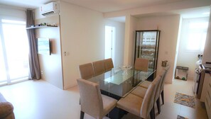 Un espace repas moderne avec une table en verre et des chaises rembourrées, un meuble vitré contenant des verres à vin, et un coin cuisine équipé d’une bouilloire et d’un évier.