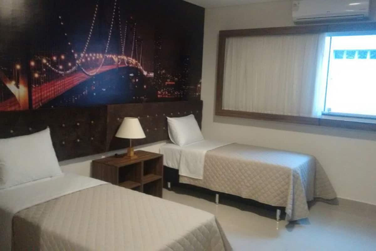 Ein Hotelzimmer mit zwei Betten, einem Nachttisch, einer Lampe, einem großflächigen Stadtbild-Wandgemälde und einem Fenster mit Vorhängen.