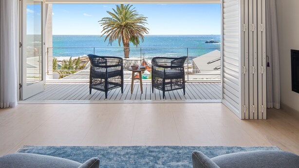 A balcony with a view of the ocean, two wicker chairs, a small table with a magazine, and a blue rug. - 52 Victoria (Cape Town)