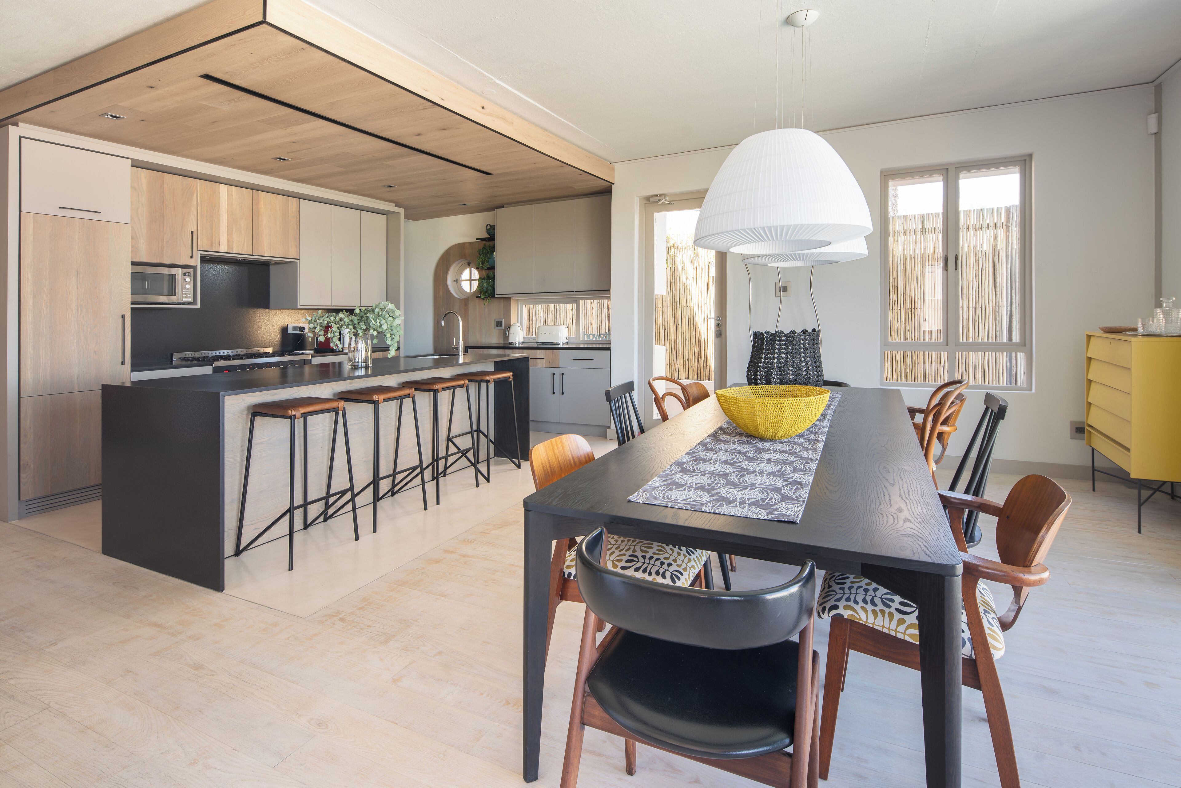 Uma cozinha moderna com ilha central, armários de madeira e espaço de refeições com uma mesa comprida e cadeiras.