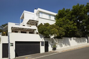 A modern two-story building with a garage, a balcony with a thatched roof, and a large tree in front. - Bayflowers Guesthouse (Cape Town)