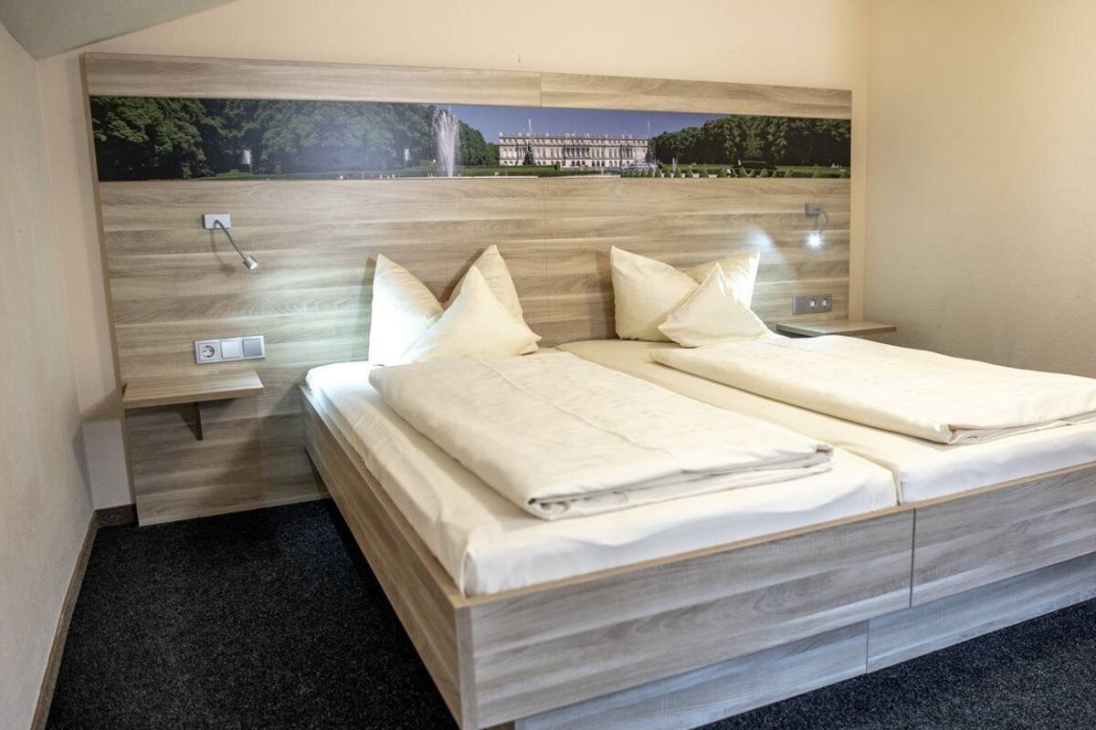 A double bed with a wooden headboard and a nightstand, a wall-mounted light, and a framed picture of a building.
