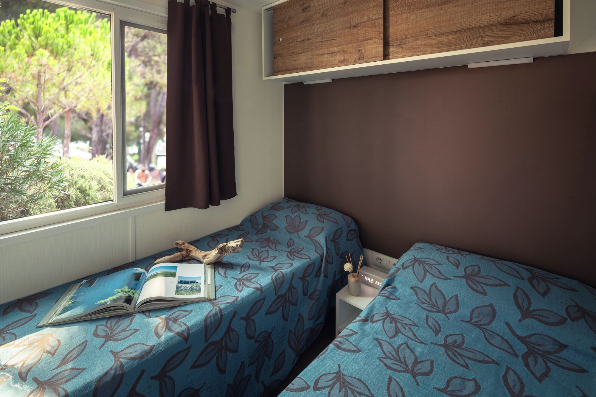 A bedroom with two beds, a window with curtains, a small table with a book and a clock, and a wooden cabinet above the beds.
