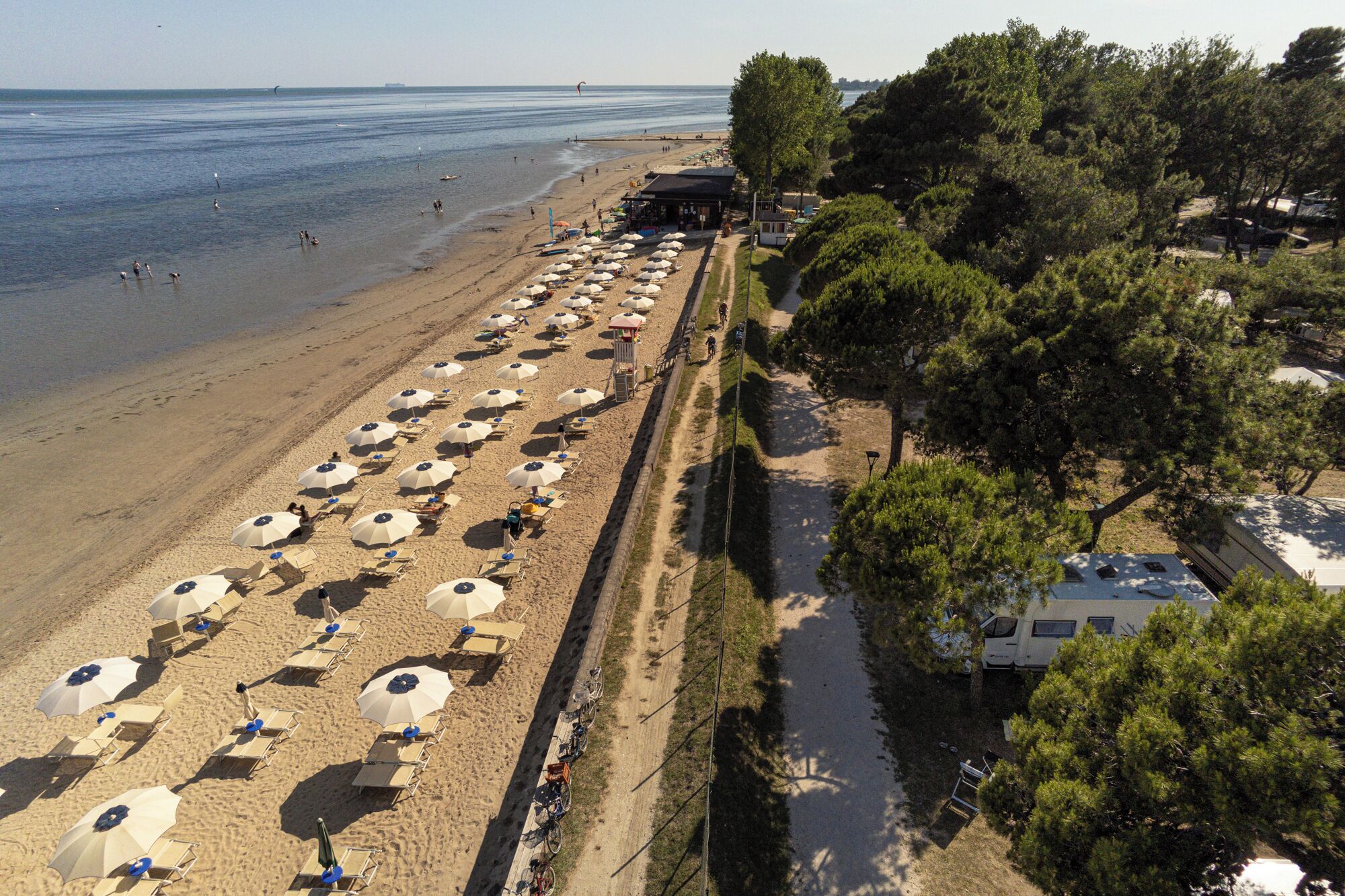 Una playa con filas de tumbonas y sombrillas, un camino bordeando la orilla, y una hilera de árboles con una caravana estacionada a la derecha.