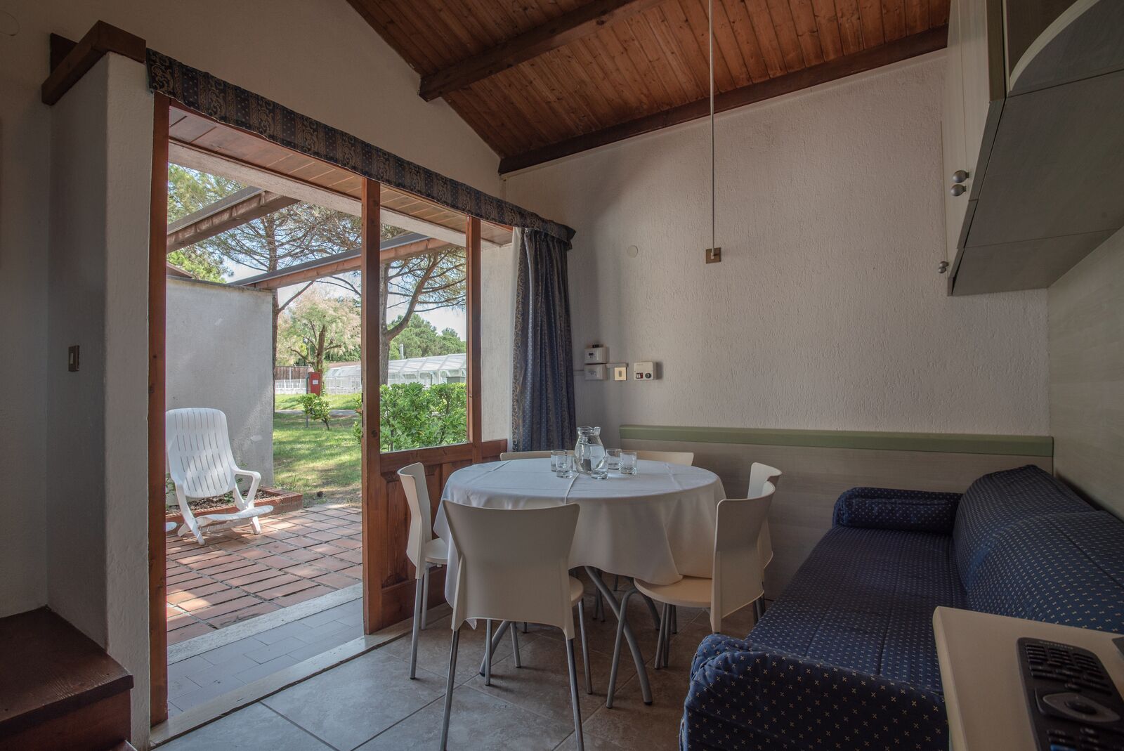 A room with a round table, white chairs, a blue sofa, and a view of a garden through a glass door.